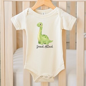 Op de afbeelding: Een crèmekleurige romper met korte mouwen en een groene aquarelillustratie van een dinosaurus met een lange nek. Onder de dinosaurus staat de tekst "Snack Attack" in een zwart cursief lettertype. Het kledingstuk heeft een ronde hals.