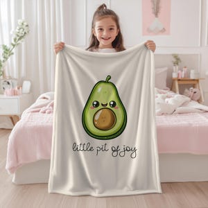 Op de afbeelding: Een crèmekleurige deken met een grote, cartoon avocado met een lachend gezicht en de tekst "little pit of joy" in zwart schrift. De avocado is groen met een bruine pit.
