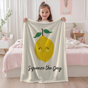 Könnte beinhalten: Cremefarbene Decke mit einem lächelnden Cartoon-Zitronen-Design. Die gelbe Zitrone hat grüne Blätter und den Schriftzug "Squeeze the Day" in schwarzer Schrift. Ein fröhliches, helles Design.