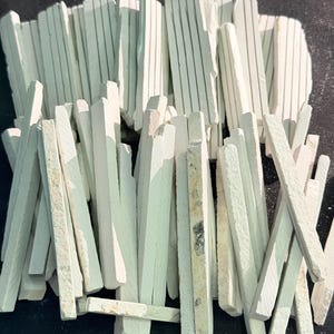 May include: A pile of white chalk sticks with thin, parallel lines. The chalk pieces are rectangular and vary in length, with some showing signs of use. The background is a dark, solid color.