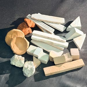 May include: A collection of natural stone pieces in diverse shapes and colours. Features rectangular, triangular, and cube-shaped stones, alongside rough, rounded rocks and a small, bowl-like object. The colour palette varies from beige and tan to reddish-brown and off-white.