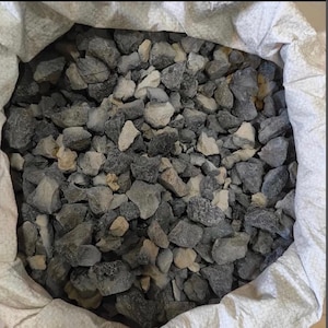 Puede incluir: Un saco tejido de color blanco lleno de una colección de piedras trituradas. Las piedras son de formas y tamaños irregulares, con una mezcla de colores gris oscuro, carbón y beige claro o blanco roto con texturas rugosas y dentadas.