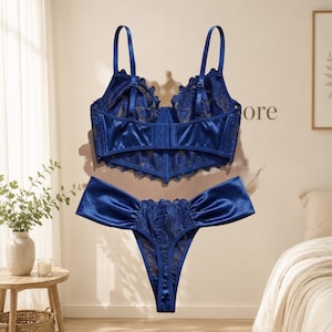 Puede incluir: Un conjunto de lencería azul real compuesto por un bustier y una tanga. El bustier cuenta con tirantes ajustables, cierre de corchetes en la espalda y un intrincado bordado floral sobre paneles de malla transparente. La tanga a juego tiene paneles laterales de satén.