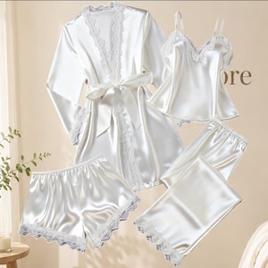 Può includere: Un set di pigiama in raso bianco composto da cinque pezzi: una vestaglia a maniche lunghe con cintura, una canotta con spalline sottili, un paio di pantaloncini, un paio di pantaloni lunghi e una gonna, rifiniti con bordo in pizzo bianco.