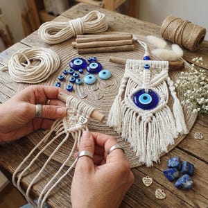 May include: A macrame wall hanging with a large blue evil eye bead, surrounded by white knotted cotton. The image also shows various blue glass beads, natural rope, and lapis lazuli stones, suggesting a craft project or handmade decor.