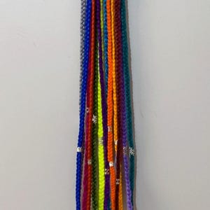 May include: A collection of colorful braided hair extensions in various shades, including blue, purple, orange, and green. The braids are adorned with small silver-colored beads. The hair extensions are hanging against a plain white wall.