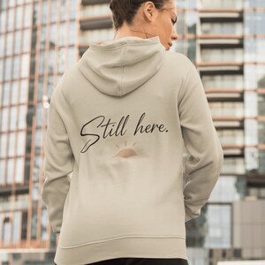 May include: A light beige hoodie with a drawstring hood. The back of the hoodie features the text "Still here." in a cursive font, with a small sun graphic underneath. The hoodie is made of a soft material.
