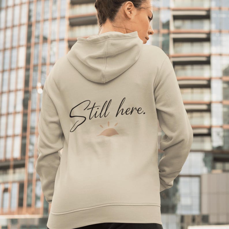 May include: A light beige hoodie with a drawstring hood. The back of the hoodie features the text "Still here." in a cursive font, with a small sun graphic underneath. The hoodie is made of a soft material.