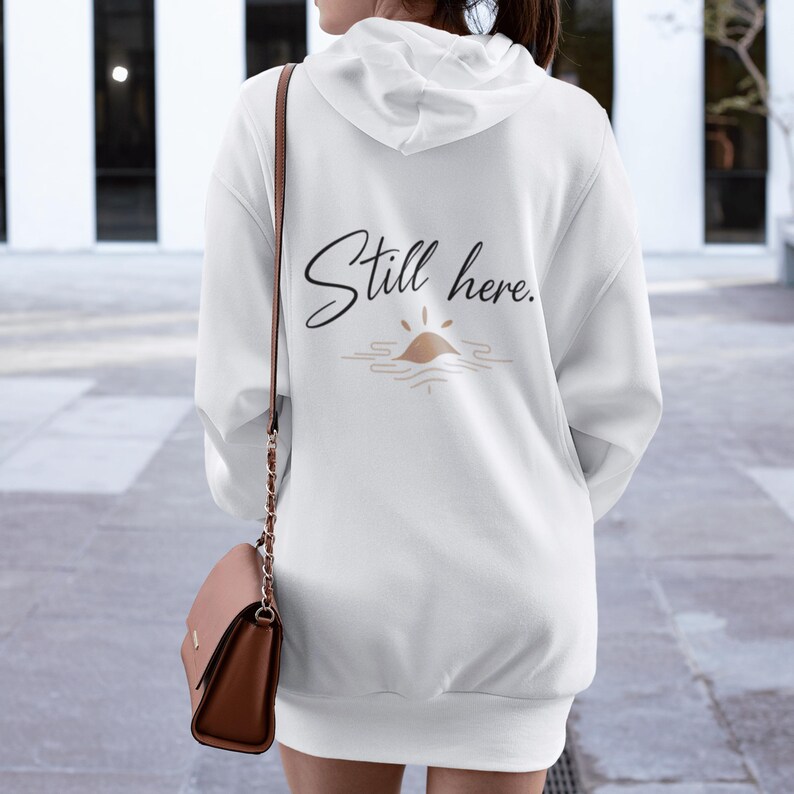 May include: White hooded sweatshirt dress with the text "Still here" in a cursive font. Below the text is a graphic of a sun over water in shades of brown and beige. The sweatshirt has a relaxed fit.
