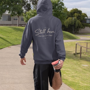 Puede incluir: Una sudadera con capucha azul marino con el texto "Still here. ONE DAY AT A TIME." en cursiva. La persona lleva pantalones cortos negros y zapatillas deportivas. La sudadera tiene capucha.