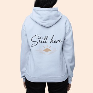 May include: A light blue hoodie with the words "Still here." in black script, above a simple sun and water graphic. The hoodie has a hood and is made of a soft material.