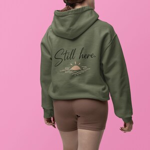 May include: Olive green hoodie with the text "Still here." in a cursive font, along with a sun and water graphic. The hoodie is made of a soft material and has a hood.