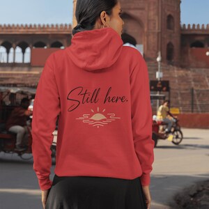 May include: A red hoodie with the text "Still here." in a script font, above a sun and water graphic. The hoodie has a hood and is being worn by a person. The background shows a building and people.