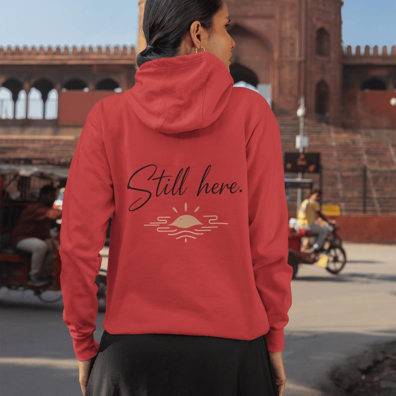 May include: A red hoodie with the text "Still here." in a script font, above a sun and water graphic. The hoodie has a hood and is being worn by a person. The background shows a building and people.