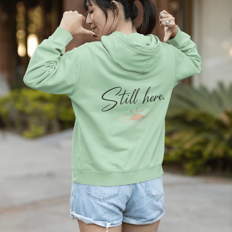 May include: A light green hoodie with the text "Still here." in black script on the back, along with a small sun graphic. The hoodie has a hood and long sleeves. The person in the image is wearing denim shorts.