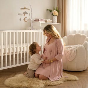 Puede incluir: Escena de guardería con una cuna blanca, un móvil con nubes y lunas, y una persona embarazada con un vestido y una bata rosas. Un niño arrodillado sobre una alfombra de piel de oveja mirando hacia arriba. Una silla acogedora y flores completan la escena.