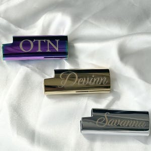 以下が含まれることがあります： カスタム刻印が施された3つの長方形の金属製ライターケース。一番上のケースは虹色の紫色で「OTN」の文字。中央のケースは金色で筆記体の「Devinn」という名前。一番下のケースは銀色で筆記体の「Savanna」という名前が刻まれています。
