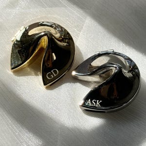 Könnte beinhalten: Zwei metallische glückskeksförmige Objekte mit polierter Oberfläche. Eines ist goldfarben mit der Gravur GD auf der Vorderseite, das andere ist silberfarben mit der Gravur ASK auf der Vorderseite. Beide verfügen über ein aufklappbares Design.