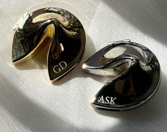 Könnte beinhalten: Zwei metallische glückskeksförmige Objekte mit polierter Oberfläche. Eines ist goldfarben mit der Gravur GD auf der Vorderseite, das andere ist silberfarben mit der Gravur ASK auf der Vorderseite. Beide verfügen über ein aufklappbares Design.