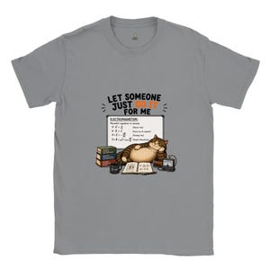 May include: A grey short-sleeve t-shirt featuring a graphic of a brown tabby cat lounging in front of a whiteboard with Maxwell's equations. The text reads: LET SOMEONE JUST DO IT FOR ME. The design includes books, a mug, and scientific equipment.