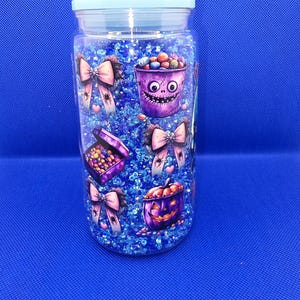 May include: A clear jar with a light blue lid filled with blue glitter and Halloween-themed decorations. The jar features illustrations of purple buckets, boxes, and bows with spiders and hearts, all filled with colorful candies.