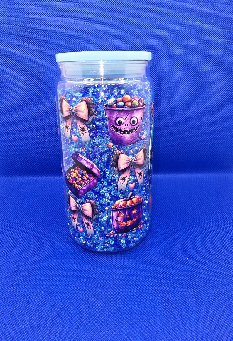 May include: A clear jar with a light blue lid filled with blue glitter and Halloween-themed decorations. The jar features illustrations of purple buckets, boxes, and bows with spiders and hearts, all filled with colorful candies.