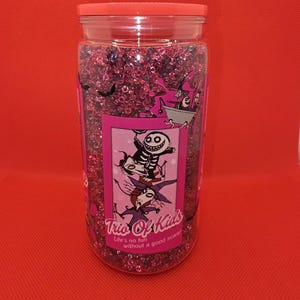 K&ouml;nnte beinhalten: Ein klares Plastikglas gef&uuml;llt mit rosa Glitzer. Das Glas hat einen rosa Deckel und ein Etikett mit Cartoon-Figuren und dem Text "Trio Of Kids". Der Text "Life's no fun without a good scare!" steht ebenfalls auf dem Etikett. Der Hintergrund ist rot.