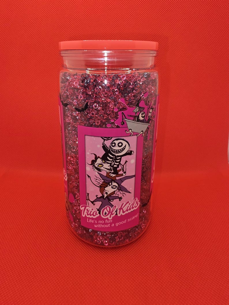 K&ouml;nnte beinhalten: Ein klares Plastikglas gef&uuml;llt mit rosa Glitzer. Das Glas hat einen rosa Deckel und ein Etikett mit Cartoon-Figuren und dem Text "Trio Of Kids". Der Text "Life's no fun without a good scare!" steht ebenfalls auf dem Etikett. Der Hintergrund ist rot.