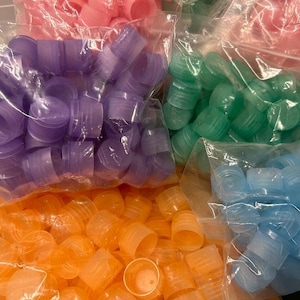 May include: Four bags of plastic bottle caps in different colors: purple, green, blue, and orange. The bags are clear plastic and the caps are arranged in a random pattern.