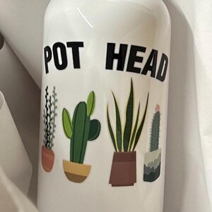 May include: White ceramic water bottle with the text "POT HEAD" in black lettering. The bottle features a design of four potted plants, including a cactus, a succulent, and two types of ferns.