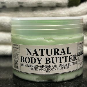 May include: A jar of natural body butter with a light green hue. The label reads "NATURAL BODY BUTTER" with "WITH MANGO~ARGAN OIL~SHEA BUTTER HAND AND BODY BUTTER". The jar is 5 oz.