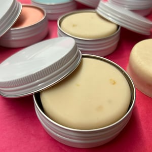 May include: A close-up of a white tin with a lid, containing a round, off-white solid product. The tin is sitting on a pink surface, surrounded by other similar tins.