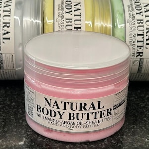 May include: A round plastic jar of pink natural body butter with a white lid and a label reading Natural Body Butter with Mango, Argan Oil, and Shea Butter, Hand and Body Butter, 5oz. Behind it are three similar jars in white, yellow, and light green.