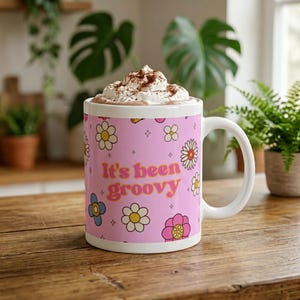 Könnte beinhalten: Eine weiße Keramik-Tasse mit rosa Hintergrund und dem Text "It's been groovy" in Gelb und Orange. Die Tasse ist mit bunten Blumenillustrationen verziert. Die Tasse ist mit einem heißen Getränk gefüllt und mit Schlagsahne und Kakaopulver bedeckt.