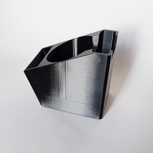 May include: A black, geometric desk tidy with multiple compartments. The organiser has a slanted design with a cylindrical holder and several rectangular sections. The surface has a textured appearance, set against a white background.