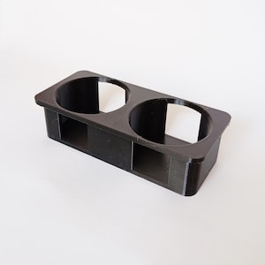 Puede incluir: Un portavasos rectangular negro con dos aberturas circulares. El soporte está hecho de un material sólido y tiene una superficie texturizada. El diseño incluye una pequeña abertura rectangular en la parte delantera.