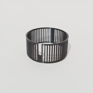 May include: A black, circular object with vertical bars creating a cage-like design. It features a partial opening on one side. The design is minimalist and modern, emphasising the geometric pattern.