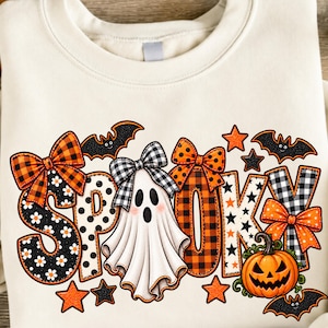 May include: A cream-colored crewneck sweatshirt featuring a graphic print of the word SPOOKY. The letters are decorated with orange and black plaid, polka dots, and daisy patterns. A white ghost with a black and white plaid bow, bats, stars, and a jack-o'-lantern are included in the design.