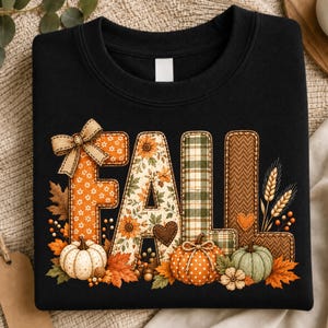 Puede incluir: Una sudadera negra de cuello redondo con la palabra FALL en letras grandes y decorativas. Cada letra tiene un patrón único que incluye flores naranjas, hojas de otoño, cuadros verdes y espiga marrón, decorado con calabazas y un lazo.
