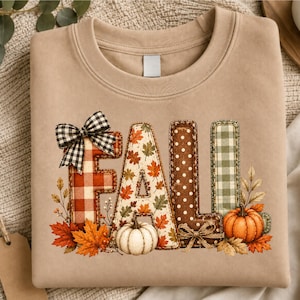 Op de afbeelding: Een beige sweater met ronde hals en het woord FALL in grote blokletters. Elke letter heeft een uniek patroon, waaronder rood ruitjespatroon, herfstbladeren, bruine stippen en groen vichy-ruitje, aangevuld met afbeeldingen van pompoenen.