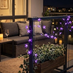 Pode incluir: Uma guirlanda decorativa de luzes com flores roxas adorna uma varanda. As luzes são enfiadas em um fio preto, com um fundo escuro de um conjunto de pátio e luzes da cidade. As flores roxas criam um efeito de iluminação ambiente.