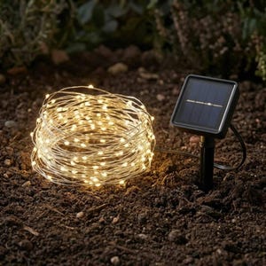 May include: A coiled string of warm white LED fairy lights connected to a black square solar panel mounted on a stake. The lights are illuminated, creating a bright glow, while the solar panel features a dark blue surface and a black frame.