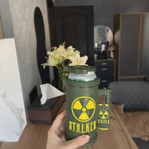 May include: An olive green cylindrical beverage can cooler featuring a yellow radiation symbol and the text STALKER printed in yellow. A small matching miniature can charm with a radiation symbol and the text STALKER hangs from the handle.