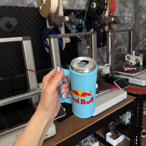 May include: A light blue Red Bull can holder with a handle. The Red Bull logo is displayed in red and yellow. The can holder is cylindrical and has a handle for easy carrying.
