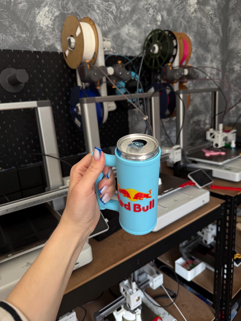 May include: A light blue Red Bull can holder with a handle. The Red Bull logo is displayed in red and yellow. The can holder is cylindrical and has a handle for easy carrying.