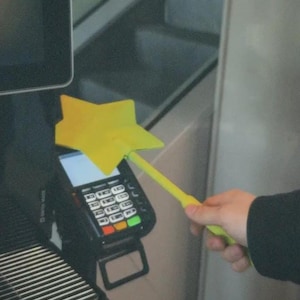 Puede incluir: Una varita mágica amarilla en forma de estrella se sostiene sobre una terminal de pago negra con una pantalla blanca y botones coloridos. La varita y la terminal están cerca de una máquina de café.