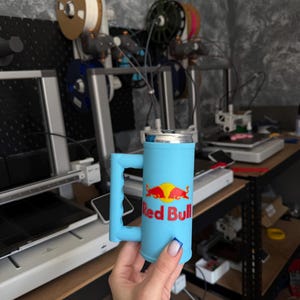 May include: A light blue 3D-printed mug with a handle, designed to hold a Red Bull can. The mug features the Red Bull logo in red and yellow. The background shows 3D printing equipment and spools of filament.