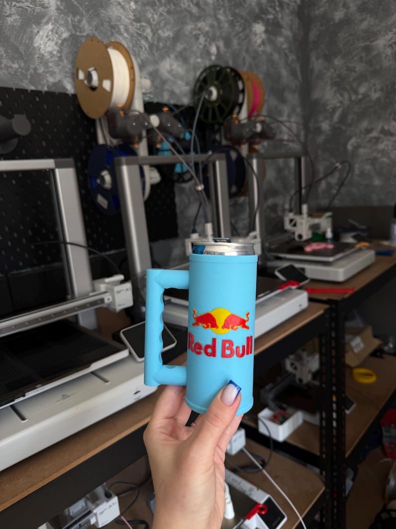 May include: A light blue 3D-printed mug with a handle, designed to hold a Red Bull can. The mug features the Red Bull logo in red and yellow. The background shows 3D printing equipment and spools of filament.