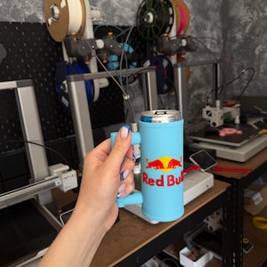 May include: A light blue Red Bull can cooler with a handle. The cooler has the Red Bull logo in red and yellow. The background shows 3D printers and spools of filament.