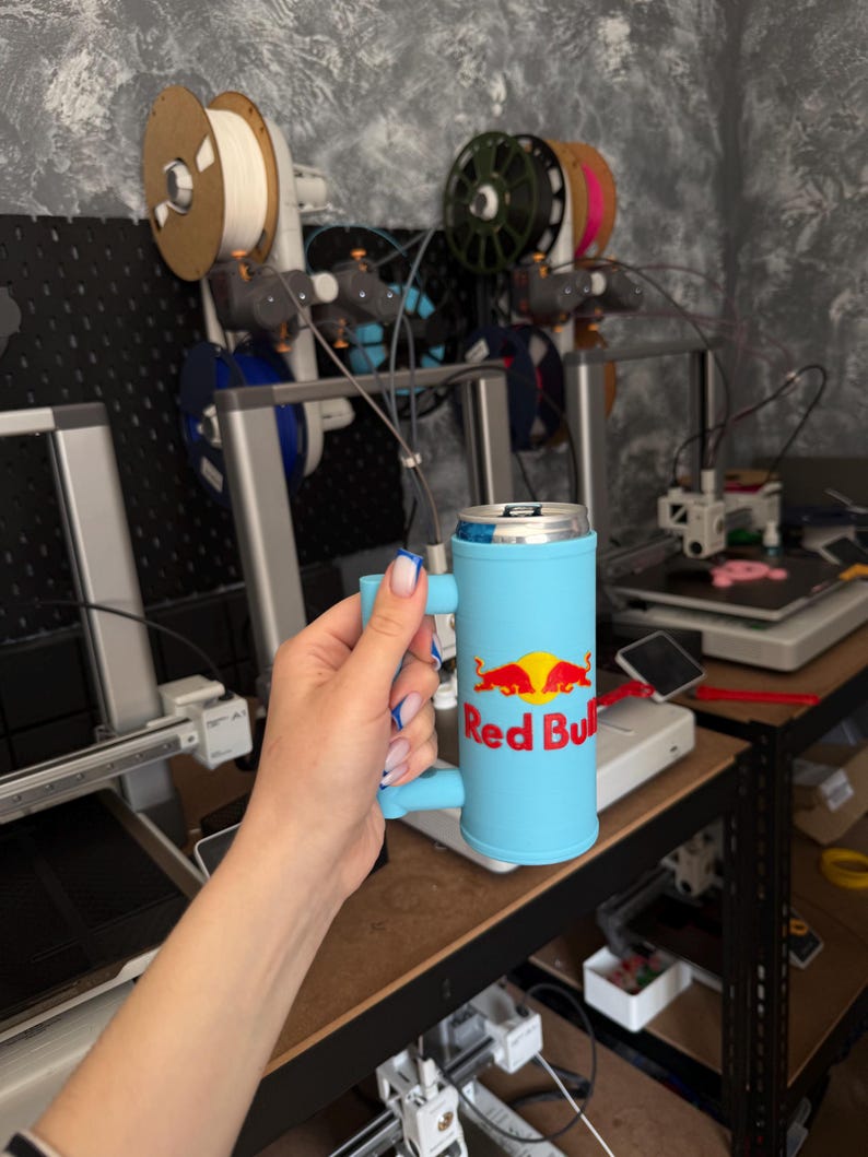 May include: A light blue Red Bull can cooler with a handle. The cooler has the Red Bull logo in red and yellow. The background shows 3D printers and spools of filament.